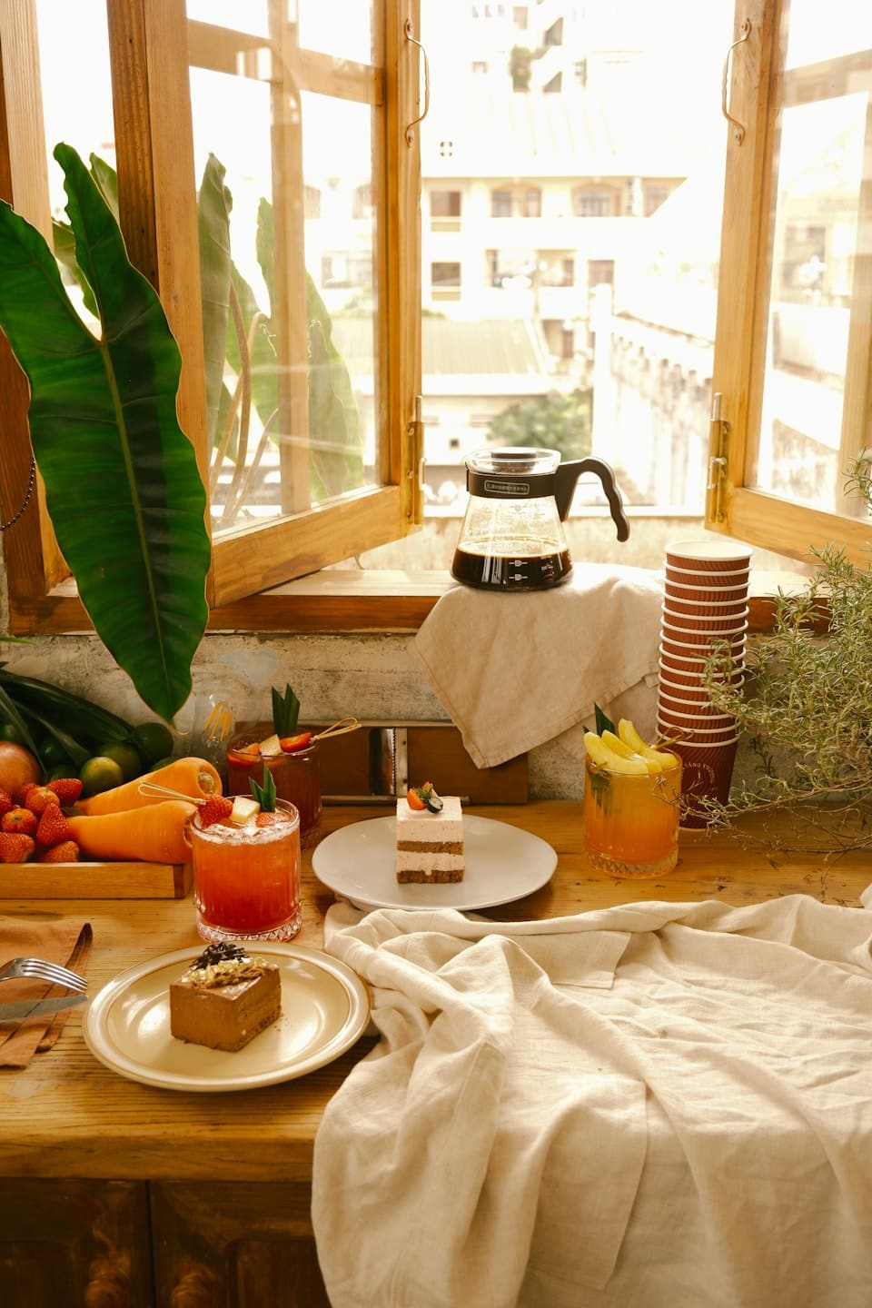 Cakes and fruit drinks sit on a table by a window.